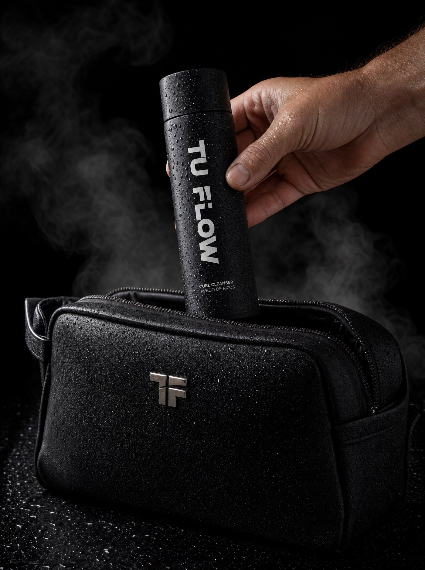Hand placing a TU FLOW Curl Cleanser bottle into a black TF toiletry bag